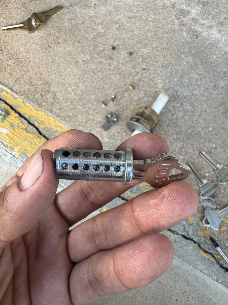 Hand holding a lock cylinder taken out of a door, with loose parts on the ground behind.